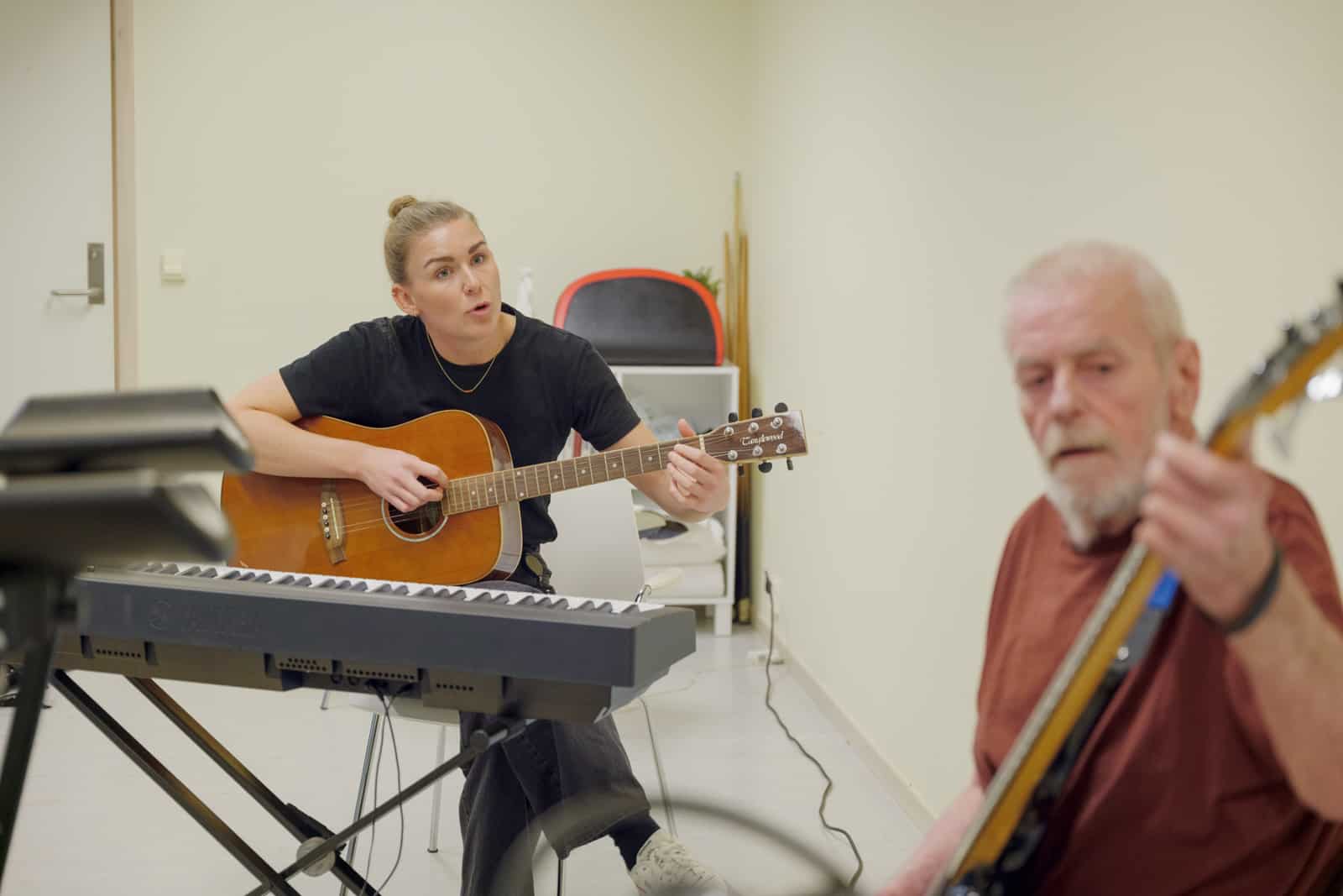 Dame som sitter foran et keyboard og spiller på en gitar mens hun synger. I forgrunnen en mann med gitar i ufokus. Fotografi.